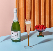 A bottle of FRE Alcohol-Removed Sparkling Brut with a filled champagne flute and a bowl of strawberries on a blue surface with a peach background and orange drape