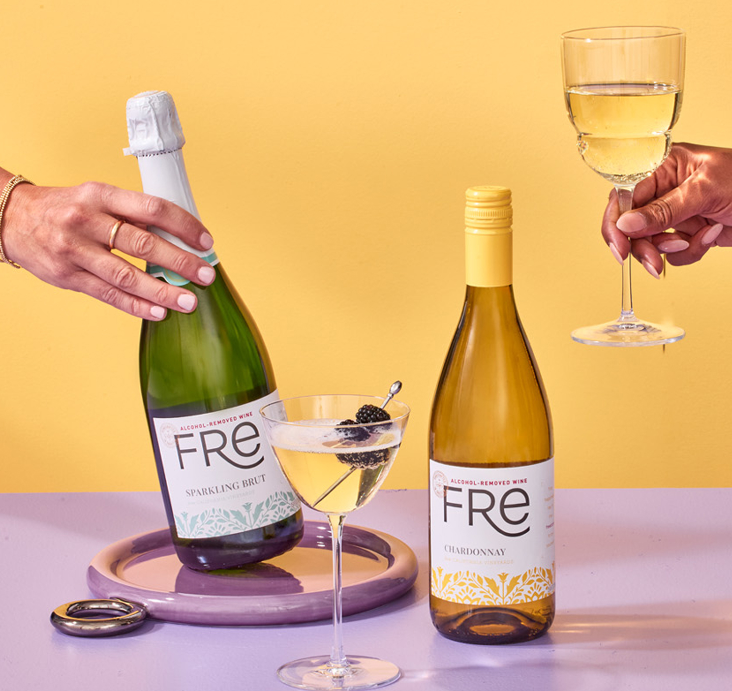 A hand grabs a bottle of Fre non-alcoholic sparkling brut wine, while another hand holds a glass of Fre Alcohol-Removed chardonnay white wine