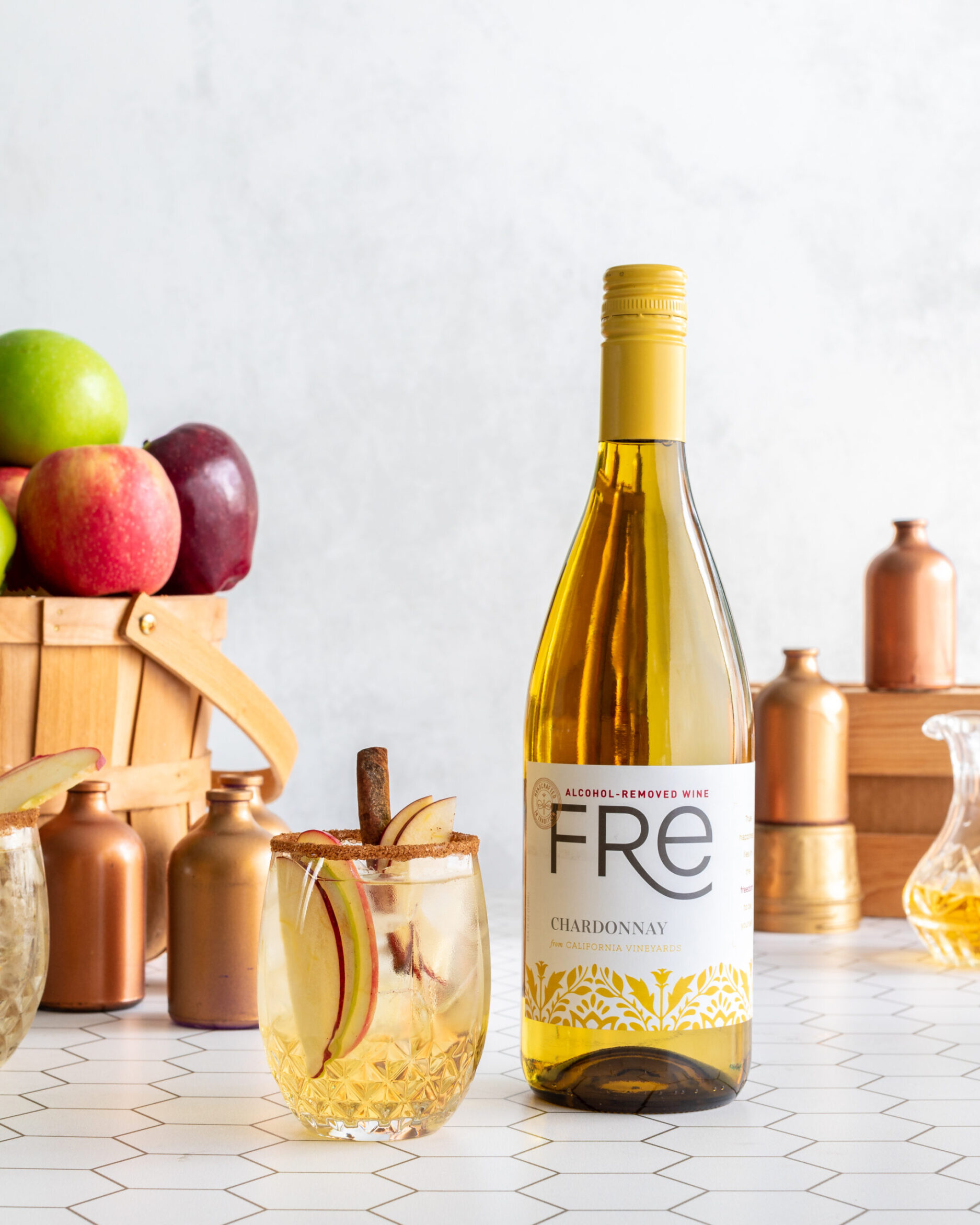 Glass of apple pie non‑alcoholic sangria with apple slices and a cinnamon stick, set in front of a bottle of FRE Alcohol‑Removed Chardonnay white wine with apples in the background.