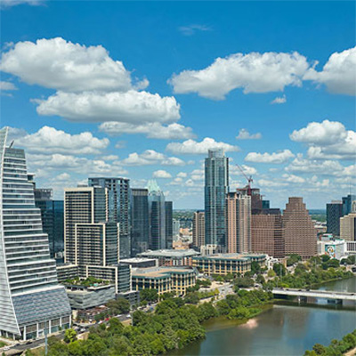Business district with multiple buildings of different heights and architectural styles, a river in the foreground, and blue sky with clouds