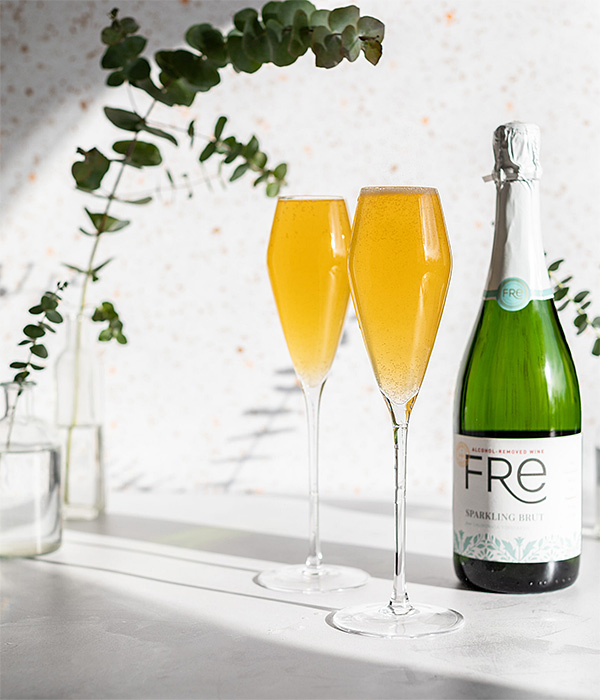 Two champagne flutes with orange-colored bellini mocktails in them, and a bottle of Fre Alcohol-Removed Sparkling brut non-alcoholic white wine