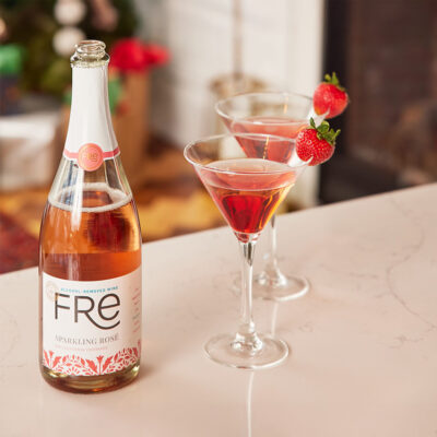 FRE Alcohol-Removed Sparkling Rose with martini glasses that contain red colored cocktail and strawberry garnish on a counter surface with a festive blurred background