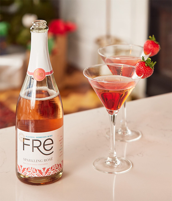 FRE Alcohol-Removed Sparkling Rose with martini glasses that contain red colored cocktail and strawberry garnish on a counter surface with a festive blurred background