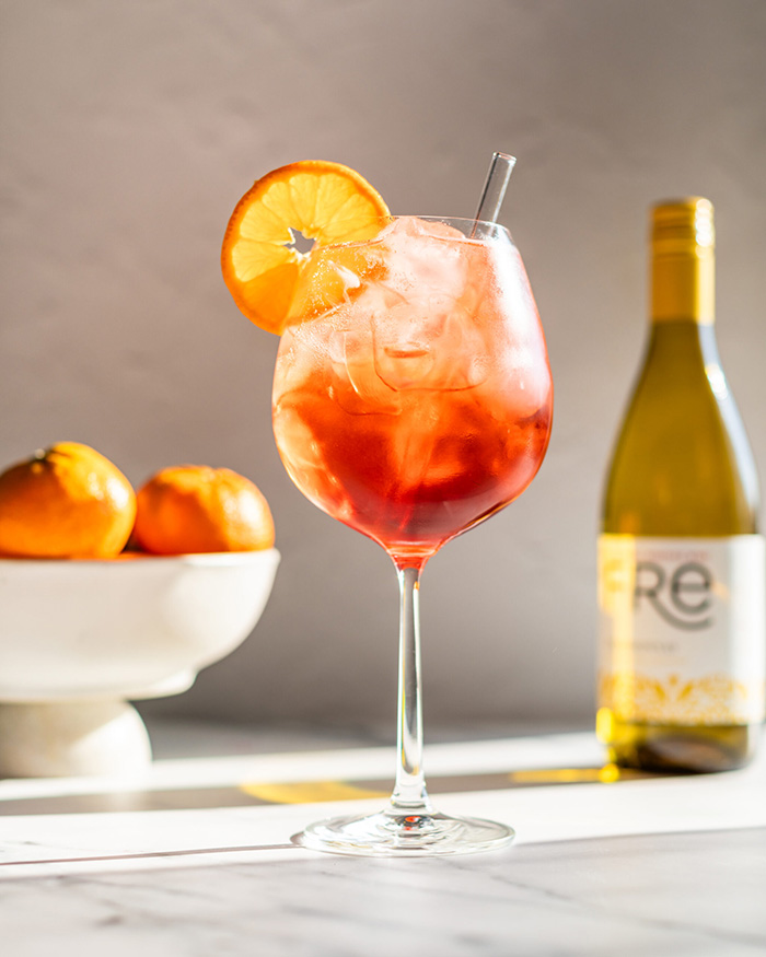 sunny image of a bright orange mocktail with a bottle of FRE Alcohol-Removed Chardonnay in the background