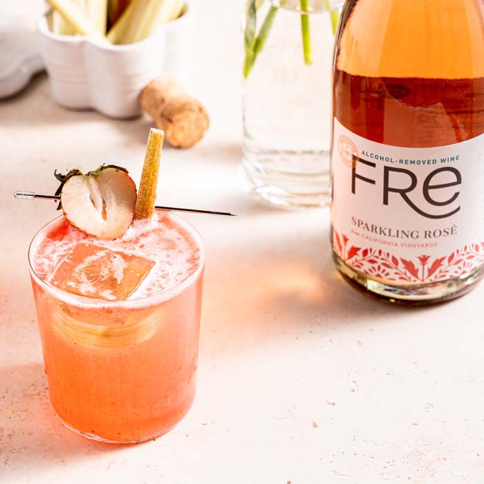 An orange colored cocktail with ice and a cut strawberry, with a bottle of FRE Alcohol-Removed Sparkling Rose, and a blurred background with a cork and various containers