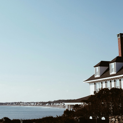 Part of a colonial house in the foreground with dormers and a chimney with a lagoon and shoreline and a large blue sky in the background