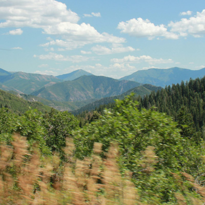 Forested hills in the distance and a blue sky with clouds with a blurred foreground indicating a photo taken from a moving vehicle