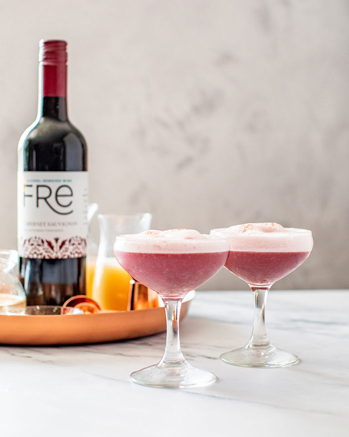 Two coupe cocktail glasses containing dusky pink apple sour mocktails with a bottle of Fre Alcohol-Removed Cabernet Sauvignon non-alcoholic red wine in the background