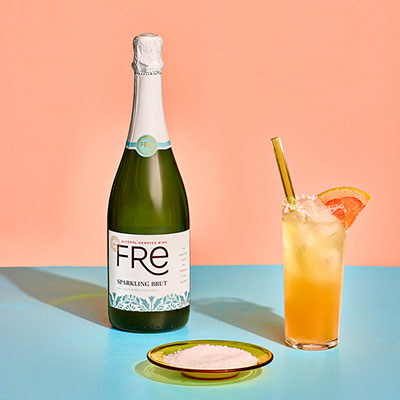 Bottle of Fre non-alcoholic sparkling brut white wine next to a saucer of salt and a summer paloma mocktail in a highball glass with a straw, garnished with a wedge of grapefruit