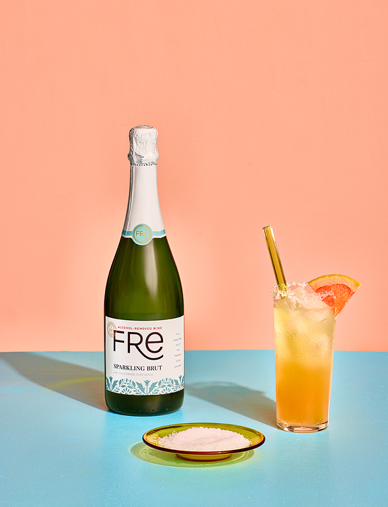 Bottle of Fre Alcohol-Removed sparkling brut white wine next to a saucer of salt and a summer paloma mocktail in a highball glass with a straw, garnished with a wedge of grapefruit