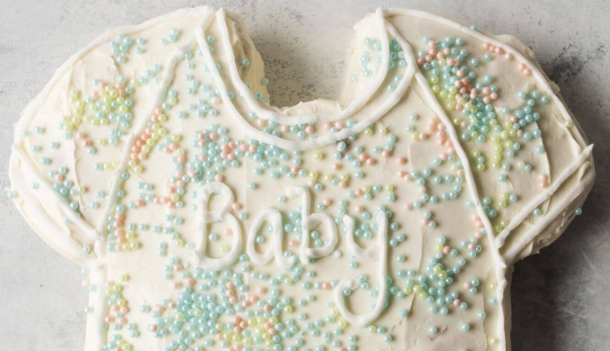 Bird's eye view of a cake shaped like a child's shirt with white frosting and pastel sprinkles with the word "Baby" written in cake frosting
