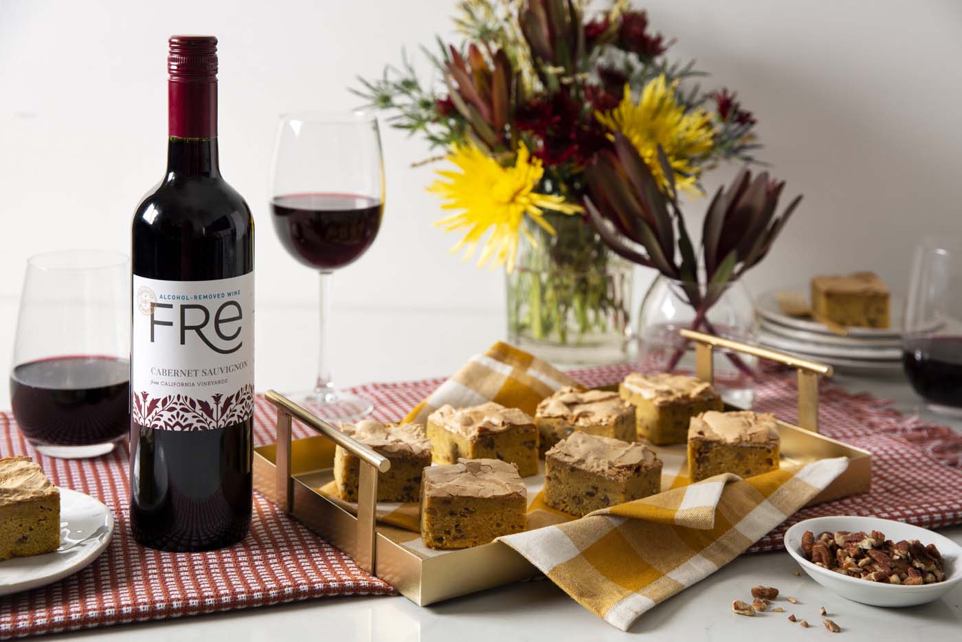 A tray of blond brownies with a bottle of FRE Alcohol-Removed Cabernet Sauvignon to the left and wine glasses filled with red wine, with a vase of flowers in the background