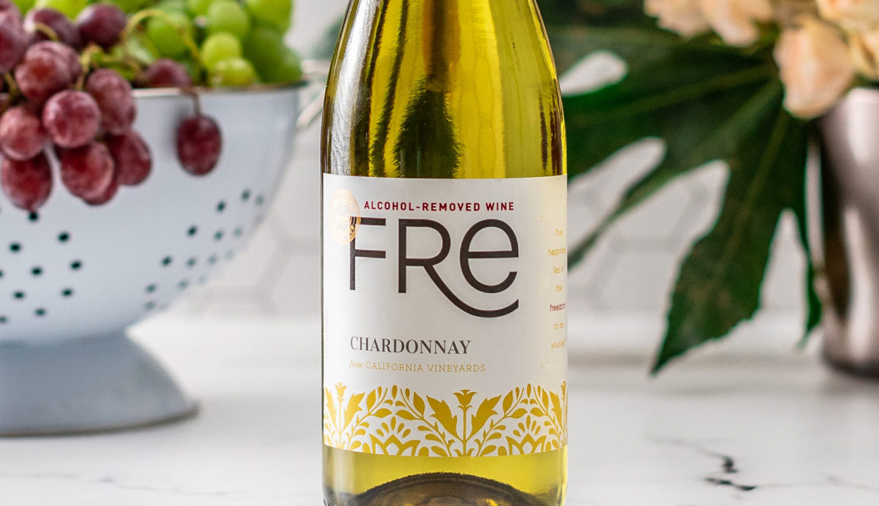 A bottle of FRE Alcohol-Removed Chardonnay in the foreground, with a blurred colander containing red and green grapes and a vase of flowers in the background