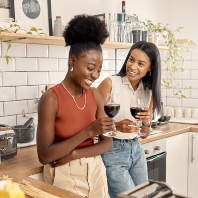 Two women standing in a stylish kitchen drinking glasses of red wine. The woman on the left appears to be laughing at something the woman on the right just said