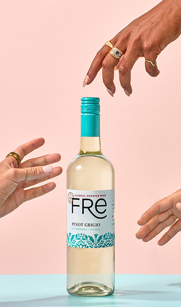 FRE Alcohol-Removed Pinot Grigio bottle on a light blue surface with a peach colored background, with three hands coming in from the sides grasping for the bottle