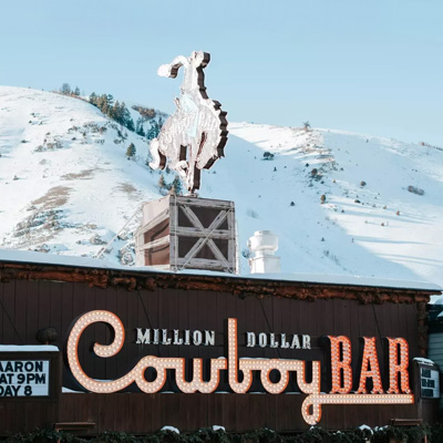 The top part of a business facade with a neon sign that reads "Million Dollar Cowboy Bar" with a bucking bronco flat sculpture on top, and snow covered hills in the background