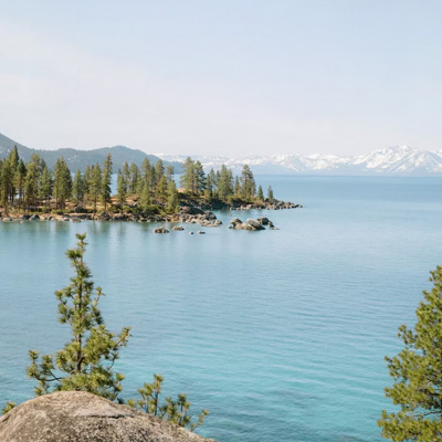 A turquoise lake with a pine covered peninsula in the left area middle ground, and snow covered mountains in the far distance