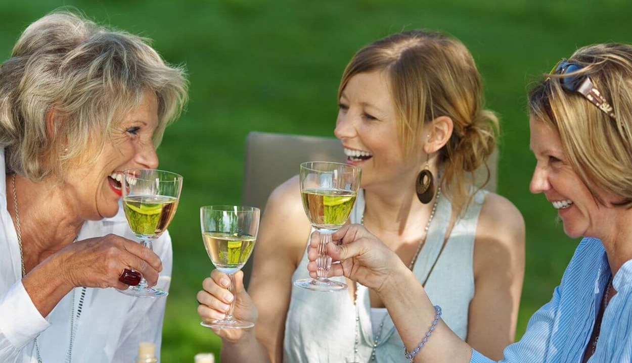 Three women in a sun-dappled outdoor setting smile at one another while they cheers glasses of white wine
