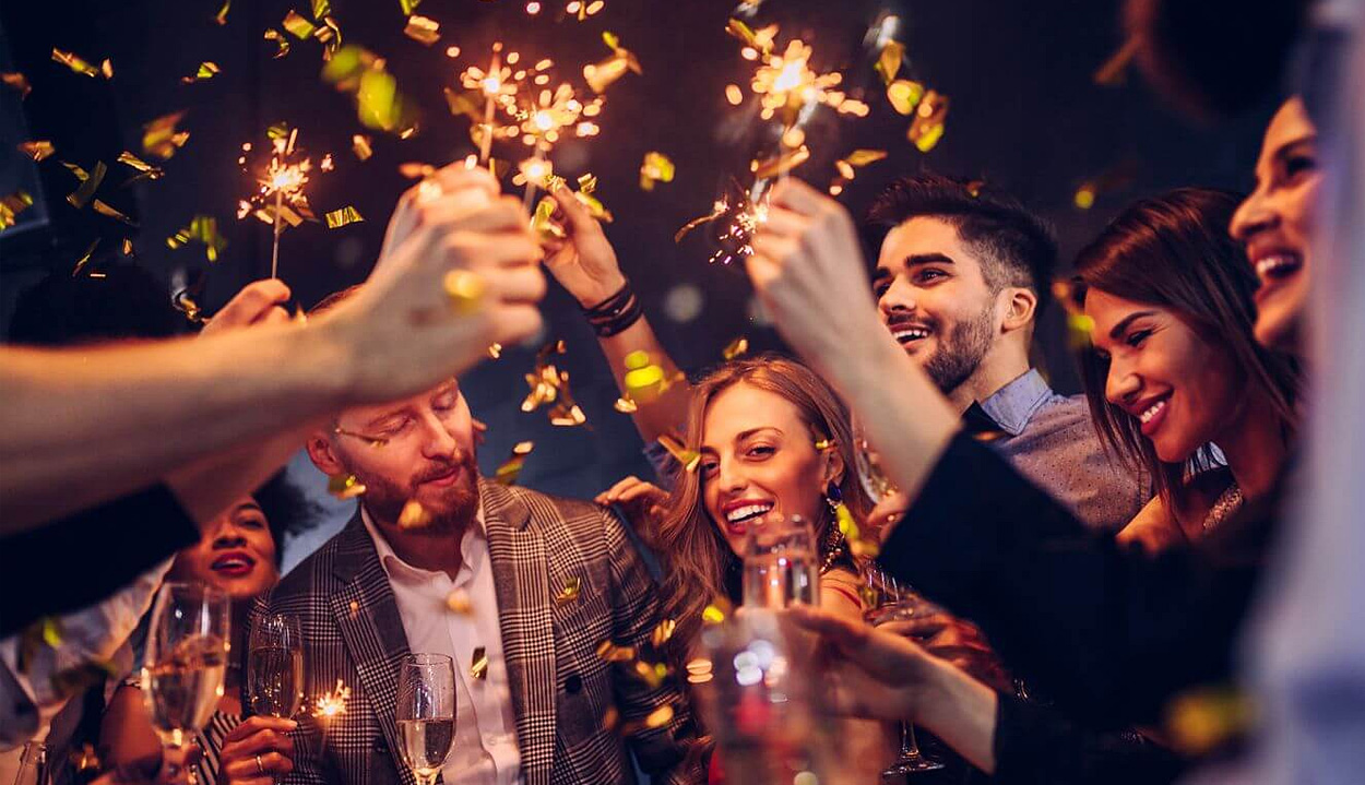 Group of well dressed people celebrating and holding up sparklers into the middle of the group and holding champagne flutes, with shiny gold confetti falling all around and a dark background