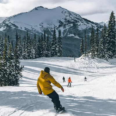 A snowboarder in the foreground headed down a slope with figures further away, and a snow covered mountain in the background