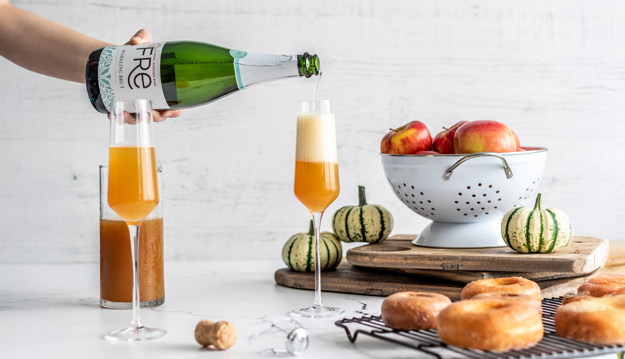 FRE Alcohol-Removed Sparkling Brut being poured into fluts with orange colored cocktails, with colander filled with apples on a cutting board, with mini pumpkins and donuts, on a marble surface