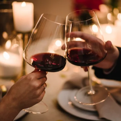 Hands holding wine glasses containing red wine clinking together for a toast, with a blurred table top in the background with candes and table settings