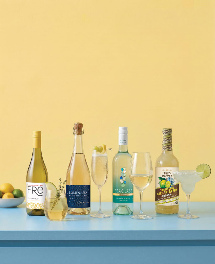 A row of four bottles of non-alcoholic FRE chardonnay, Luminara sparkling, Seaglass Sauvignon Blanc, and Tres Agaves classic lime margarita mix stand next to glasses of the same drinks