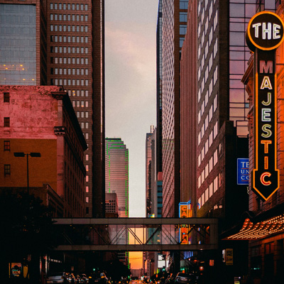 A city scene of Dallas Texas with a sunset sky in the background peeking through a street with buildings on either side and a neon sign in the foreground, with street and traffic lights in the lower foreground and a pedestrian arial bridge crossing between the buildings